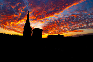 Dramatic, fiery sunset with orange cirrus clouds and silhouette of a church or cathedral spire and tower. Colourful evening sky with sun rays and majestic clouds with silhouetted town skyline. . - Powered by Adobe