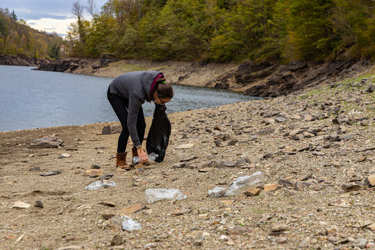 Young female picks up litter and plastic bottles from a beauty spot; girl collects rubbish to help the community; woman bending over to gather plastic waste from a lake shore in a national park.