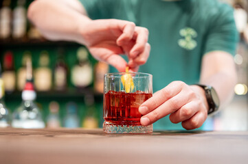 Person in green shirt garnishing a red cocktail with citrus twist at a bar, with blurred bottles in the background