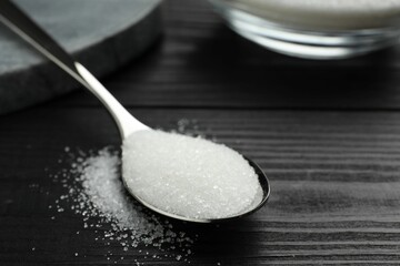 Spoon with granulated sugar on black wooden table, closeup