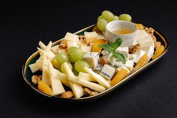 Cheese platter with grapes, nuts, dried apricots, and honey on black background