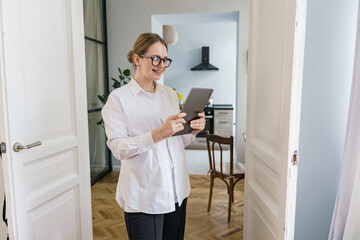 A contented woman with glasses uses a tablet, standing by a door in a stylish apartment.