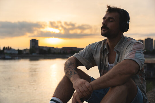 Young hipster millennial man sitting on riverside looking at city listening music on wireless headphones enjoying moment, life and sunset. Happy independent male enjoy freedom and life concept