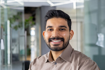 Portrait of a cheerful young man wearing a light brown shirt, smiling confidently in a contemporary office setting. Perfect image for business and lifestyle themes.