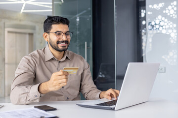 Smiling young businessman using laptop and credit card for online shopping. He is sitting in a...