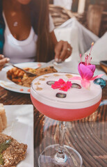 A glass of pink drink with a flower on top sits on a table next to a plate, Food and drink