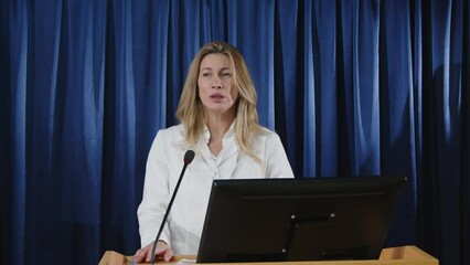 Female health official standing by lectern on stage and speaking in microphone in front of journalists during press conference
