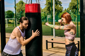 A woman in sportswear engage in a boxing workout in a park, guided by a personal trainer,...
