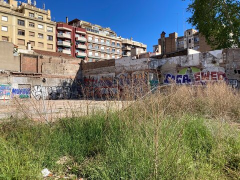 Solar abandonado en Barcelona, con viviendas al fondo. Muros con graffitis. Día soleado. Parcela vacía.