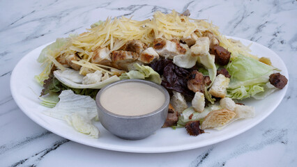 Caesar salad. Closeup view of fresh a salad with lettuce, parmesan cheese, bread croutons, sliced grilled chicken breast, bacon and Caesar dressing on the white marble table