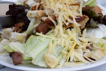 Caesar salad. Closeup view of fresh a salad with lettuce, parmesan cheese, bread croutons, sliced grilled chicken breast, bacon and Caesar dressing on the white marble table with a wooden background	