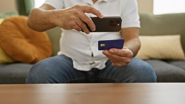 Mature man photographs credit card with smartphone at home, showing security and technology in finance.
