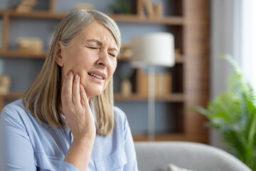 An elderly Caucasian woman sits at home, touching her cheek and grimacing in pain. She appears to be suffering from a severe toothache or jaw discomfort. - obrazy, fototapety, plakaty