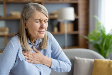 Mature woman experiencing discomfort and holding her chest due to pain. She seems anxious and...
