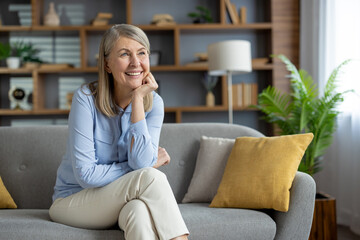 A cheerful senior woman sits comfortably on a gray sofa at home, looking contemplative and smiling,...