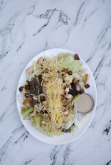 Caesar salad. Top view of fresh a salad with lettuce, parmesan cheese, bread croutons, sliced grilled chicken breast, bacon and Caesar dressing on the white marble table.