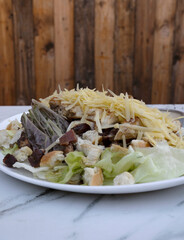 Caesar salad. Closeup view of fresh a salad with lettuce, parmesan cheese, bread croutons, sliced grilled chicken breast, bacon and Caesar dressing on the white marble table with a wooden background	
