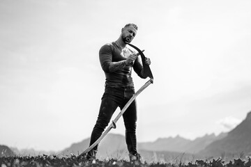 Strong farmer using scythe. Sexy Farmer with a scythe on green grass field. Muscular Farmer cut...