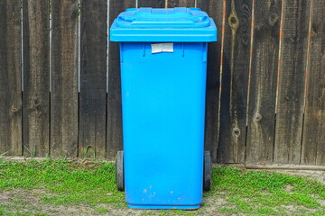 one plastic blue closed trash bin on wheels stands on the ground during the day near a wooden fence on the street