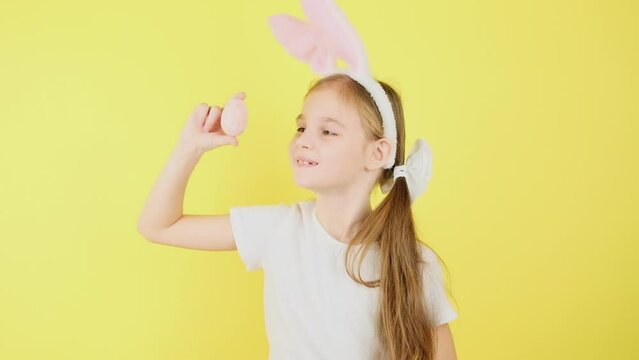Portrait child in bunny ears, looking at camera. Cute little girl pointing on colored egg, posing isolated over yellow color background.