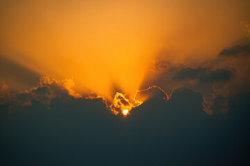 The sun is setting behind a cloud. The sky is orange and the clouds are dark. The sun is shining...