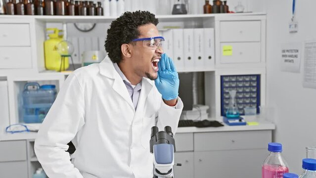 Bewildered young african american man scientist wearing safety glasses, arms wide, clueless in the lab with a 'no idea' expression.