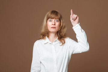 Portrait of attractive Middle-Aged Woman with Blonde Hair Pointing finger at up, showing promo, looking at camera, standing against beige background
