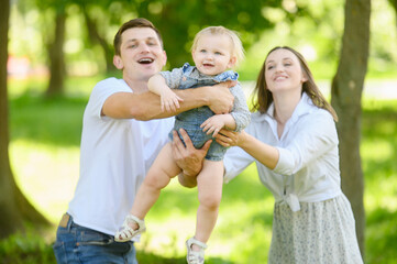 Family is having fun outdoors in park: mom and dad swing in arms, throw up child, laugh, feeling happy. Have fun weekend together, pleasant memories, holidays