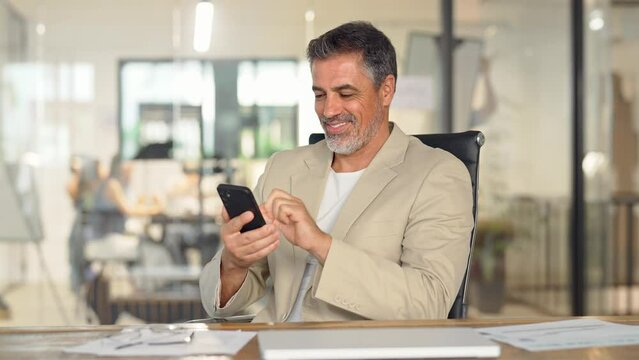 Happy busy middle aged business man ceo investor sitting in office using mobile cell phone. Mature businessman professional entrepreneur looking at smartphone at work making baking payment.
