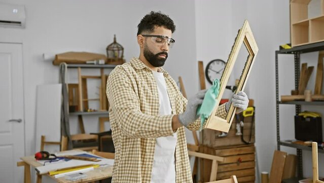 Bearded man cleaning picture frame in a woodworking workshop wearing safety goggles.