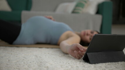 Pregnant woman laid on mat at living-room floor checking tablet device selecting prenatal exercise guidance app - obrazy, fototapety, plakaty