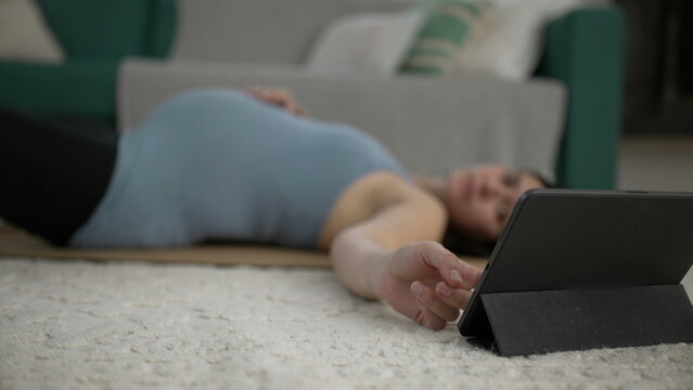 Pregnant woman laid on mat at living-room floor checking tablet device selecting prenatal exercise guidance app