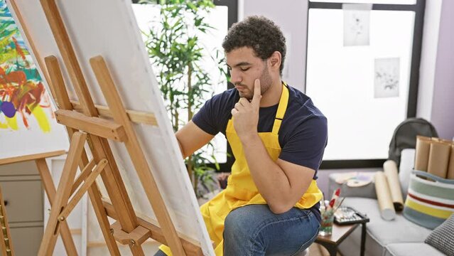 A contemplative man in a yellow apron standing indoors, assessing his own artwork on an easel with thoughtful gestures.
