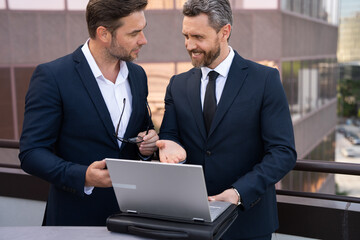 Two businessmen on the street, deeply engrossed, eyes fixed on their laptop screen, perhaps...