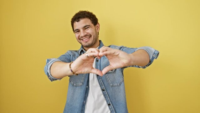Charming young arab man, joyfully standing before a yellow isolated background, fashioning a heart symbol with hands, evoking a romantic concept of love