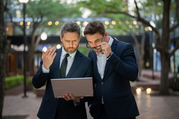 Two businessmen on the street, deeply engrossed, eyes fixed on their laptop screen, perhaps...