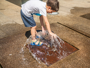 Hot summer day in the city. Little boy is having fun playing in the park fountain. He is playing barefoot getting wet and splashing.