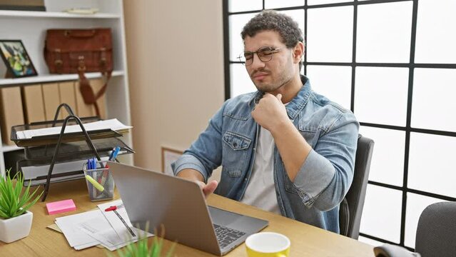 Young man with glasses and beard experiencing neck pain while working on laptop in modern office