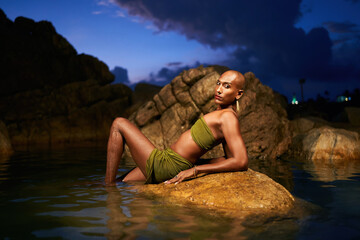 Trans sexual black fashion model in open dress, brass jewelry poses lying on big stone inside natural pool surrouded by rocks on tropical island at night. Androgynous ethnic person in backwater.