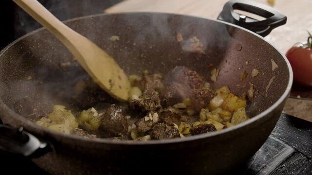 Cook carefully mixes the almost ready-made, delicious potatoes with the meat using a wooden spatula. A fragrant aroma of smoke rises from the food. Close up. Slow motion. 4k
