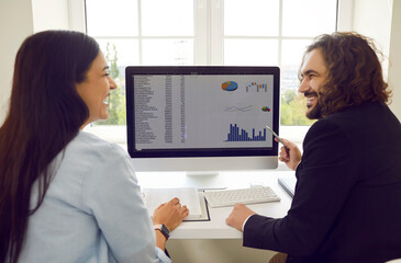 Two cheerful colleagues in office are discussing joint work project and analyzing financial graphs. Man and woman look at each other and smile while sitting in front of working computer monitor.