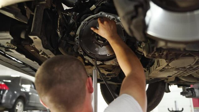 Auto mechanic installs clutch disc in car. Repairman works on vehicle gearbox, replaces release bearing, adjusts clutch plate. Workshop technician fixes transmission components under lifted auto.