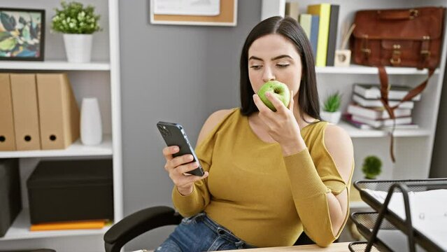 Hispanic woman in office eating apple while browsing smartphone, portraying a casual work break.