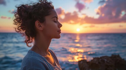Young woman inhales the salty air as the sun sets over the ocean in a blaze of orange and pink. - obrazy, fototapety, plakaty