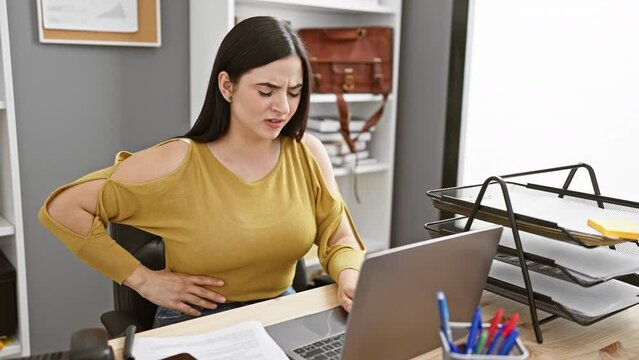 A young hispanic woman experiences stomach pain while working at her office computer, showcasing a moment of discomfort in a professional setting.
