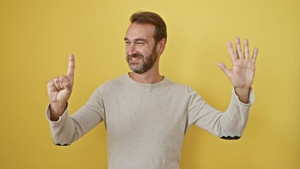 Cheerful middle-aged hispanic man with beard, standing against isolated yellow background, confidently pointing up, showing the number six with fingers, beaming a happy smile