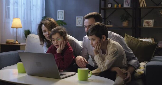 Full family using laptop computer browsing through websites, social media. Happy couple with children planning vacation, booking hotel, shopping, ordering delivery sitting on the sofa together