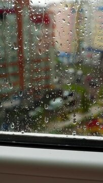 Glass window in rainy weather. Raindrops falling down on the urban background. View from the flat on the rainy weather in the city. Vertical video