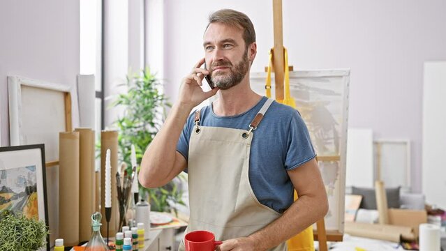 Handsome bearded man in apron chats on phone while holding red cup in an art studio filled with paintings and supplies.