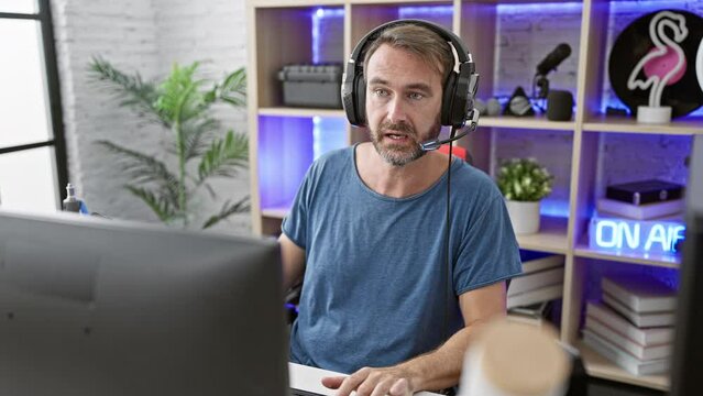 Middle-aged bearded man wearing headphones in a modern lit home office with gaming setup and 'on air' sign.
